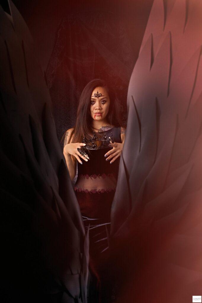 Goth woman standing in front of black wings holding a black crown waiting