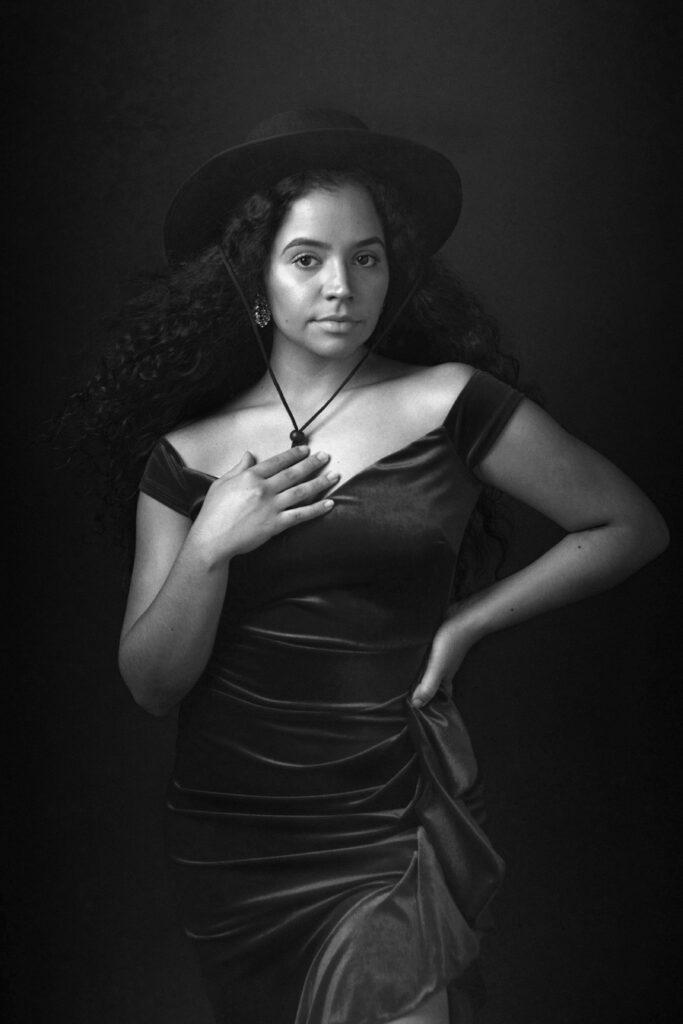 Black and white portrait of a young woman posing wearing a hat and a velvet dress